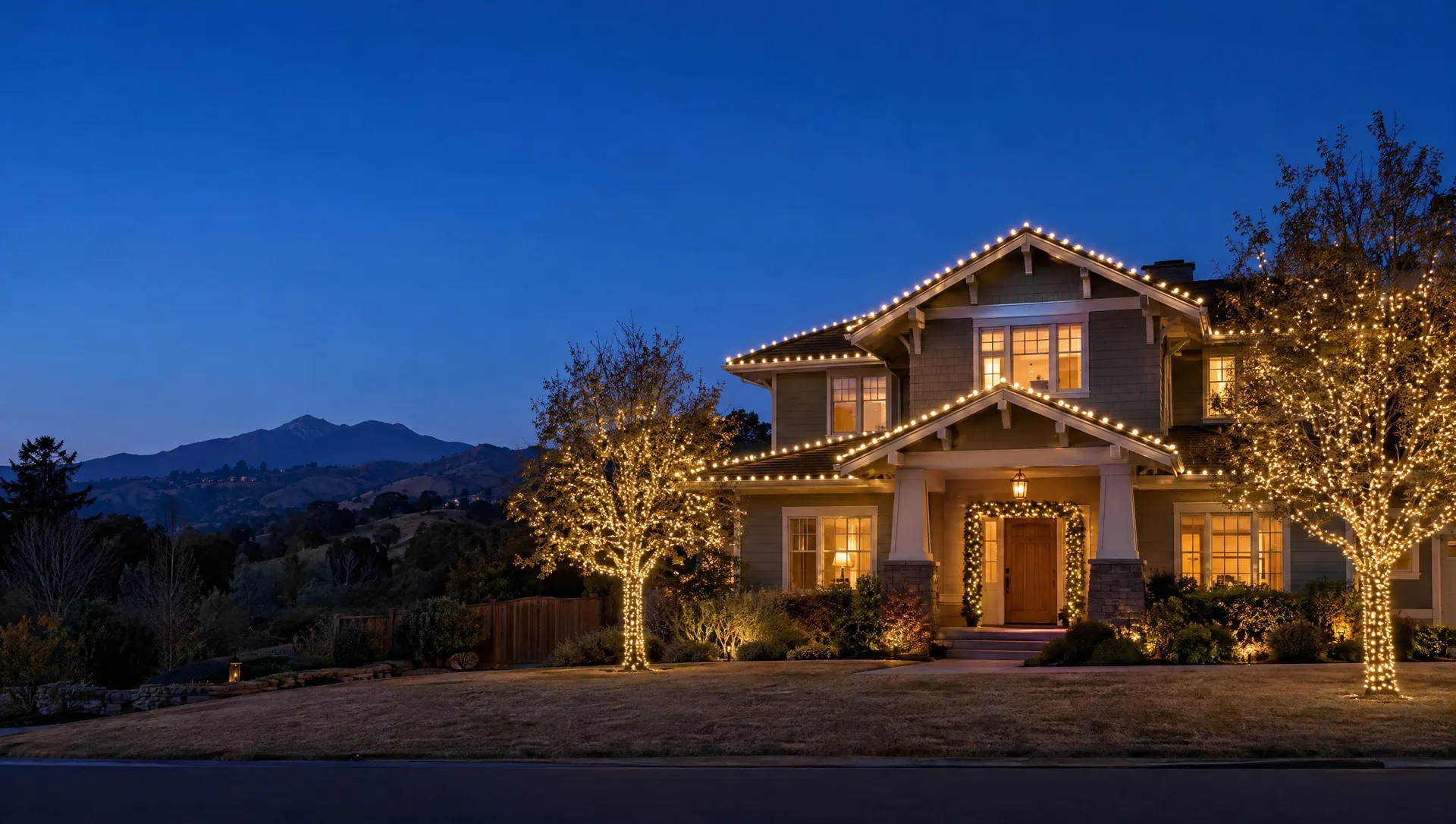 Two-story craftsman home at blue hour with warm white lights along every roofline, wrapped front-yard trees and Mount Diablo in the distance