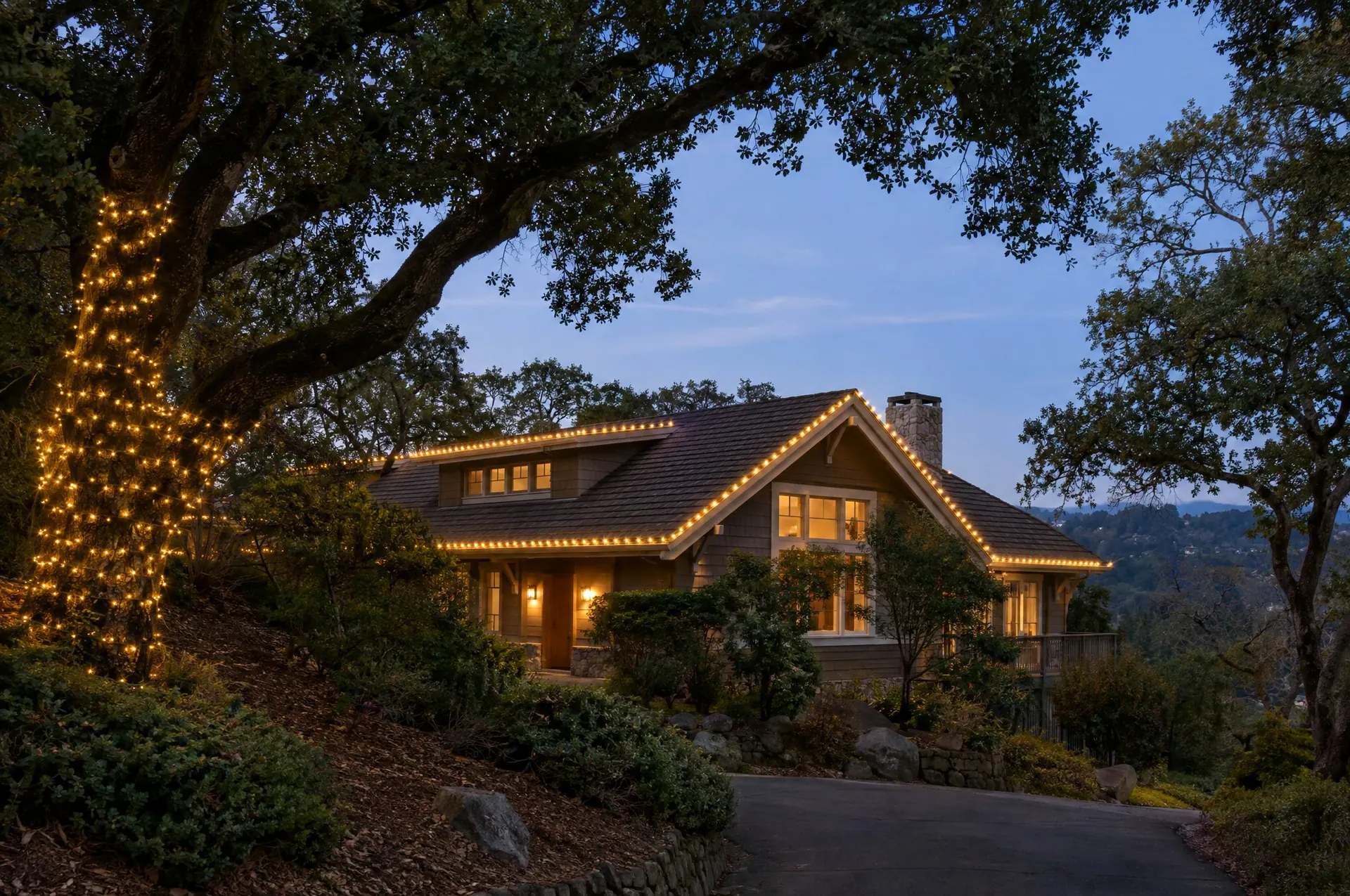 Shingle-roof home among large oak trees at dusk with warm white roofline lights and one wrapped oak trunk